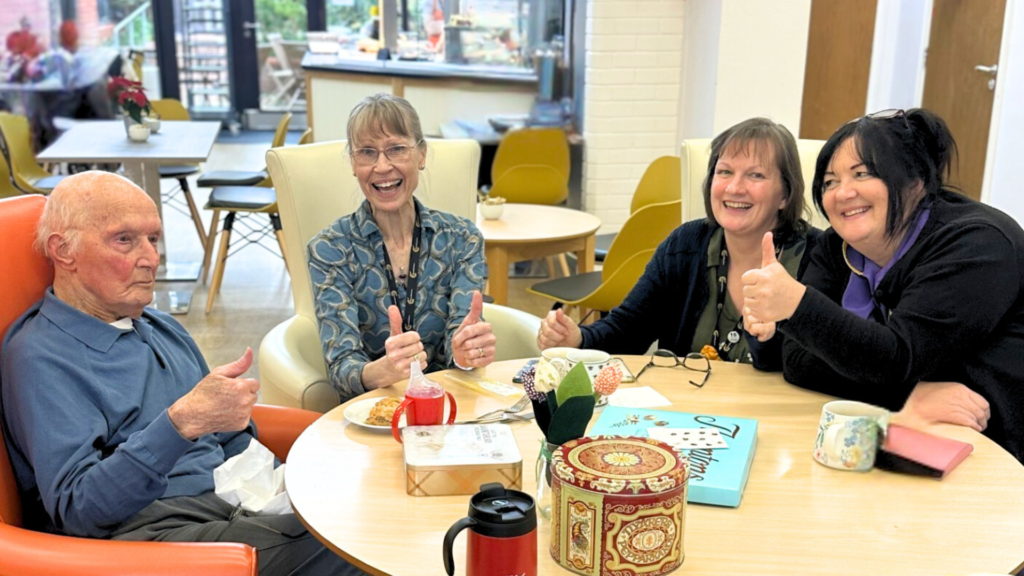 A group of people - a mix of elderly people and workers sit round a table enjoying refreshments. They are smiling and appear happy, posing for the camera with thumbs up.