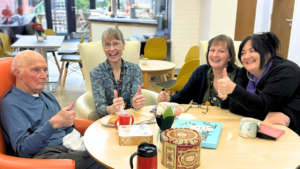 A group of people - a mix of elderly people and workers sit round a table enjoying refreshments. They are smiling and appear happy, posing for the camera with thumbs up.