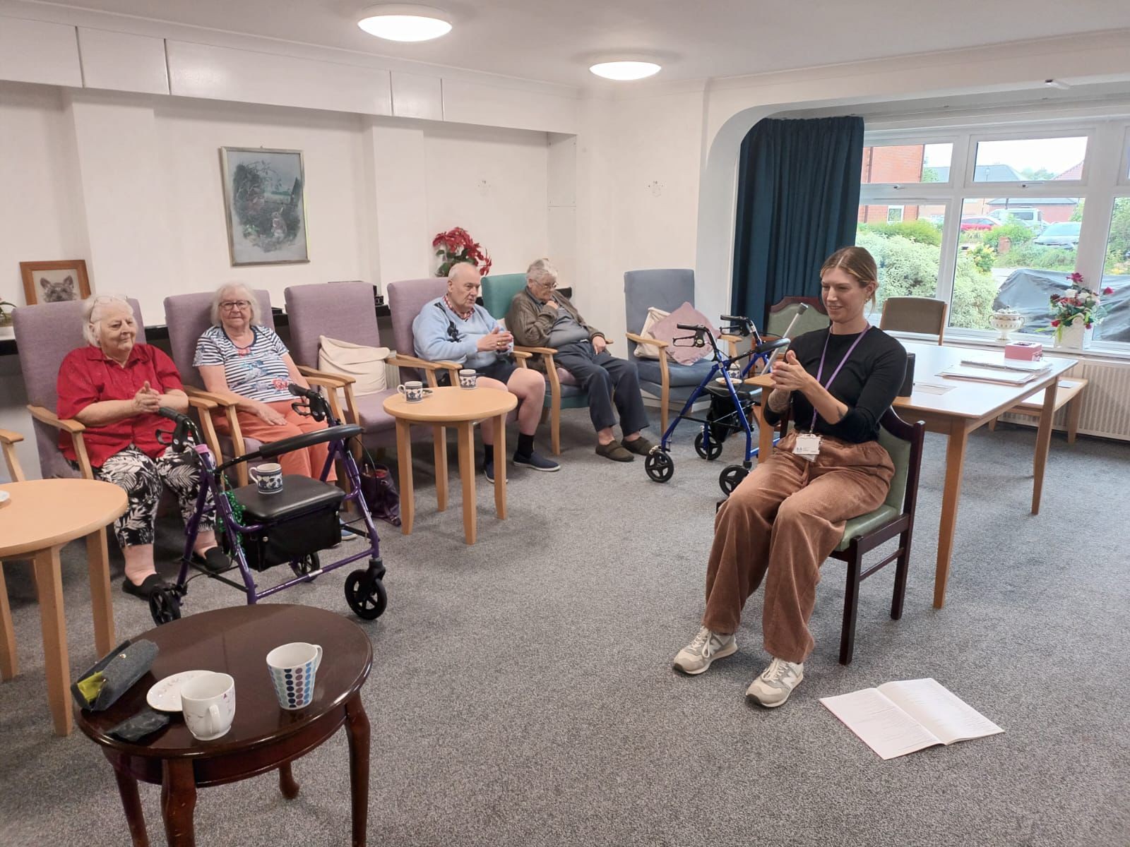 A group of older people sit in chairs following a female instructor with gentle seated exercise at age concern hampshire