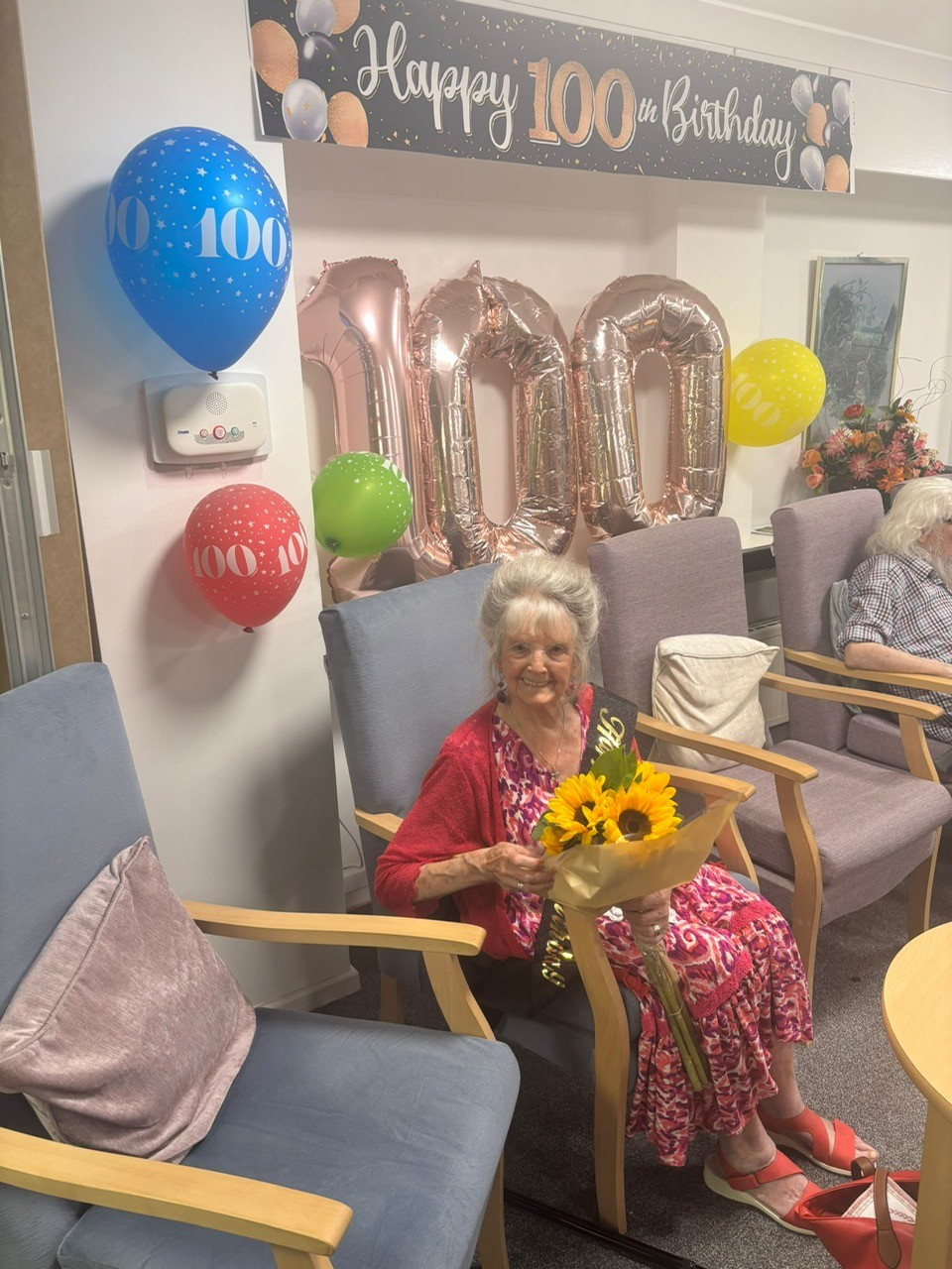 A picture of a lady in a pink dress holding sunflowers. She is sat smiling with 100th birthday balloons behind her at her 100th birthday party